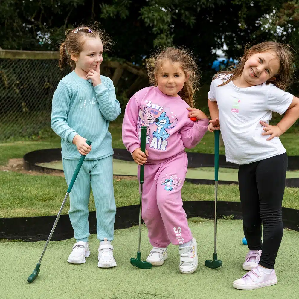 Three young girls pose playfully on the Navan mini-golf course, each holding a putter. They are dressed in colourful casual clothes and smiling, with greenery and a wire fence in the background.
