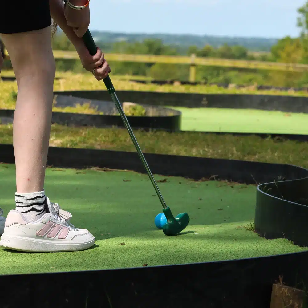A person in white trainers and striped socks prepares to hit a blue golf ball with a mini-golf club at Navan mini-golf, an inviting green outdoor course surrounded by trees and grass.