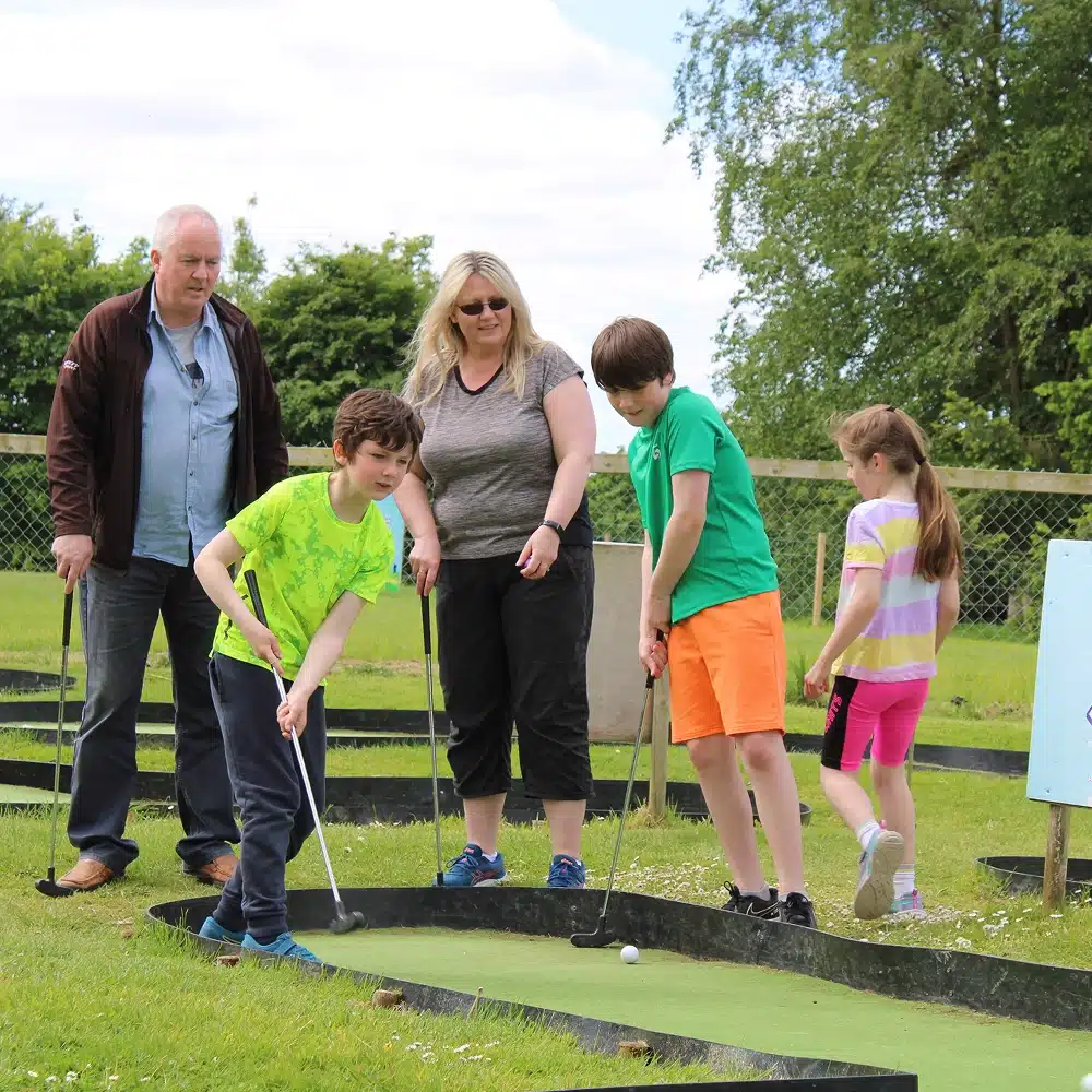 Four people, two adults and two children, watch as a boy in a green shirt prepares to hit a mini-golf ball at Navan crazy golf. Another boy and a girl stand nearby with golf clubs on the sunny, pleasant outdoor course.