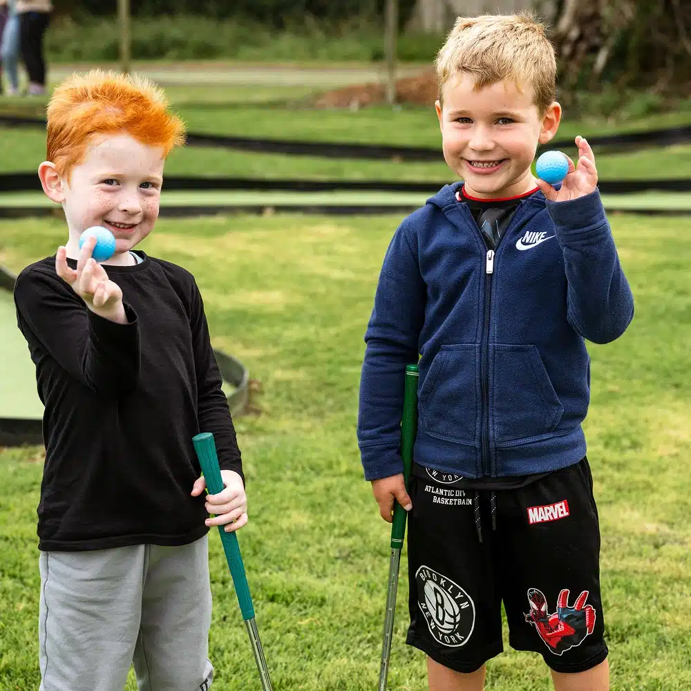 Two young boys stand on grass at Navan mini-golf, holding mini-golf clubs and blue golf balls, smiling at the camera. One wears a black shirt, the other a blue jacket and black shorts with Marvel graphics.
