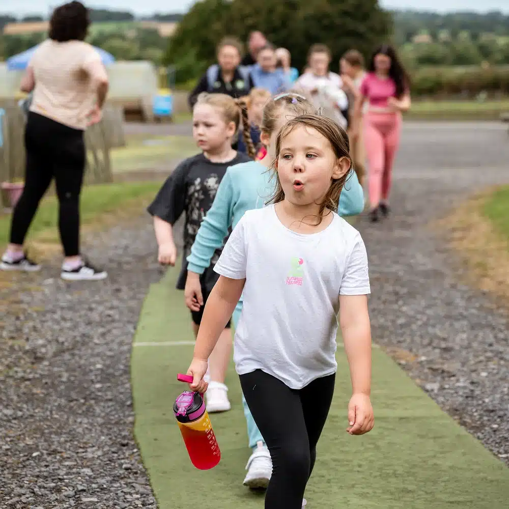 A group of children walk outdoors in a line on a green mat at Navan mini-golf, with a girl in a white shirt and black leggings holding a pink water bottle at the front. Adults and greenery are visible in the background.