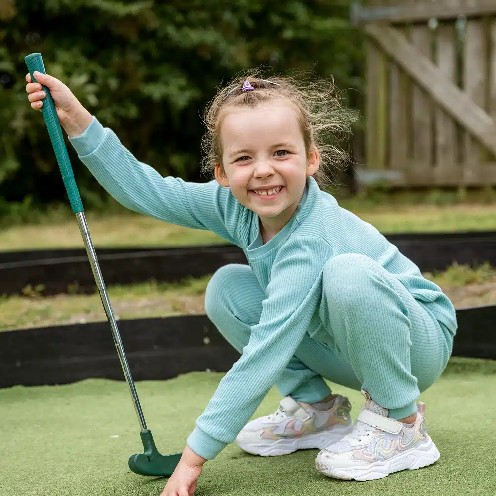 A young girl in a light blue tracksuit and colourful trainers crouches and smiles at the camera while holding a mini-golf putter and placing a ball on the Navan mini-golf putting green outdoors.