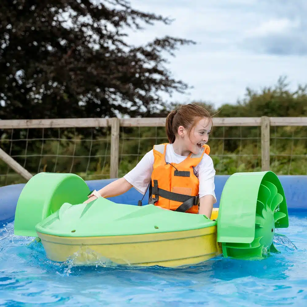 A young girl wearing an orange life jacket is smiling as she rides a small green and yellow Navan pedalo boat in a blue pool outdoors. Trees and a wooden fence are visible in the background.