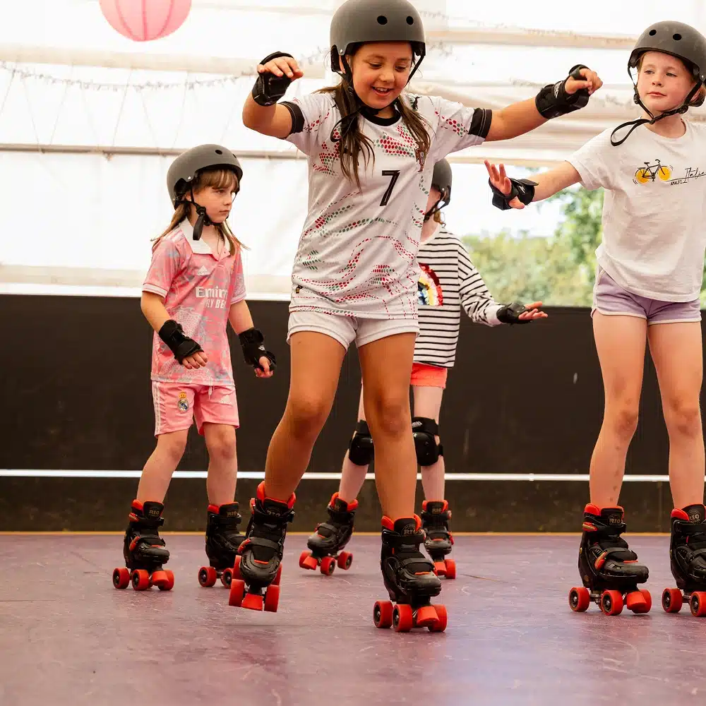Four children wearing helmets and protective gear roller skate indoors at a Navan roller disco party, focusing on balance and coordination. They appear to be having fun whilst learning to skate together.