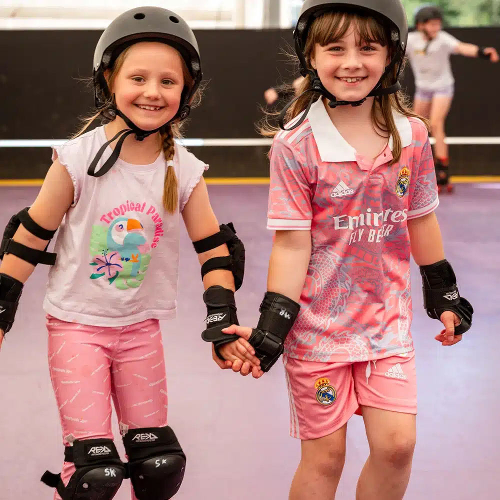 Two young girls wearing helmets and protective pads smile whilst holding hands and roller skating indoors at a Navan roller disco party. Both are dressed in pink outfits, with other skaters visible in the blurred background.