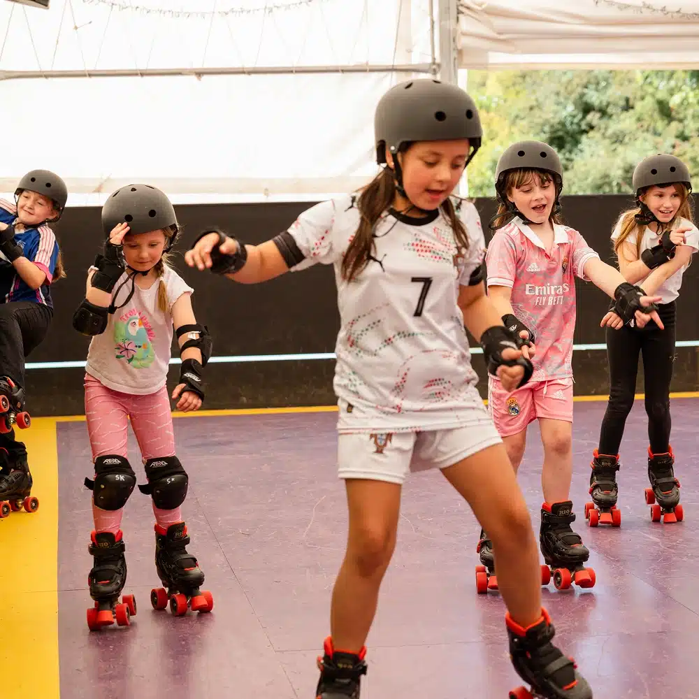 Five young children wearing helmets and protective gear roller skate indoors on a purple and yellow floor, enjoying a Navan roller disco party as they learn and practise together, some smiling and holding out their arms for balance.