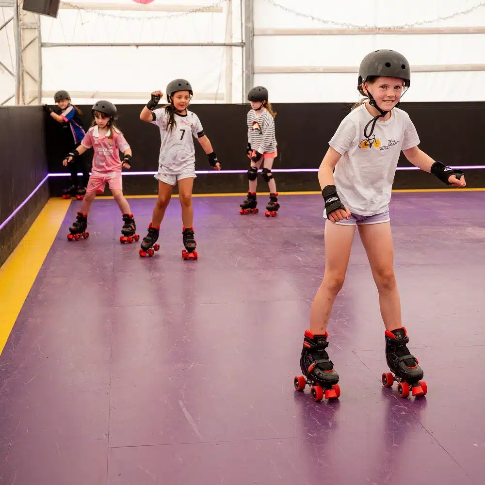 Five children wearing helmets and protective gear enjoy a Navan roller disco party, skating indoors on a purple floor. One girl in front smiles at the camera, whilst the others skate or pose in the background near the wall.