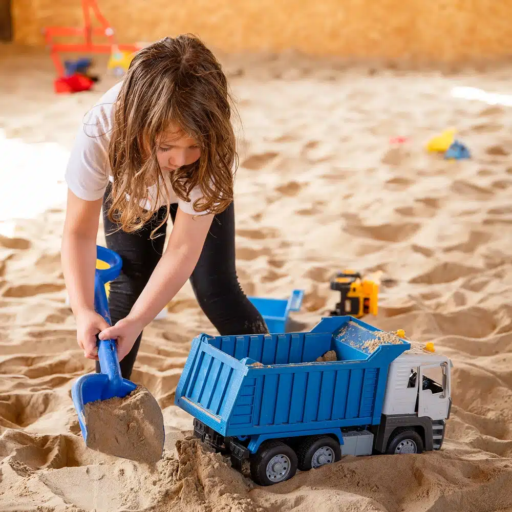 A young child with long hair uses a blue plastic spade to load sand into a large blue toy dumper lorry while enjoying Navan The Sandbarn activities in a sandy indoor play area. Toy construction vehicles are scattered in the background.