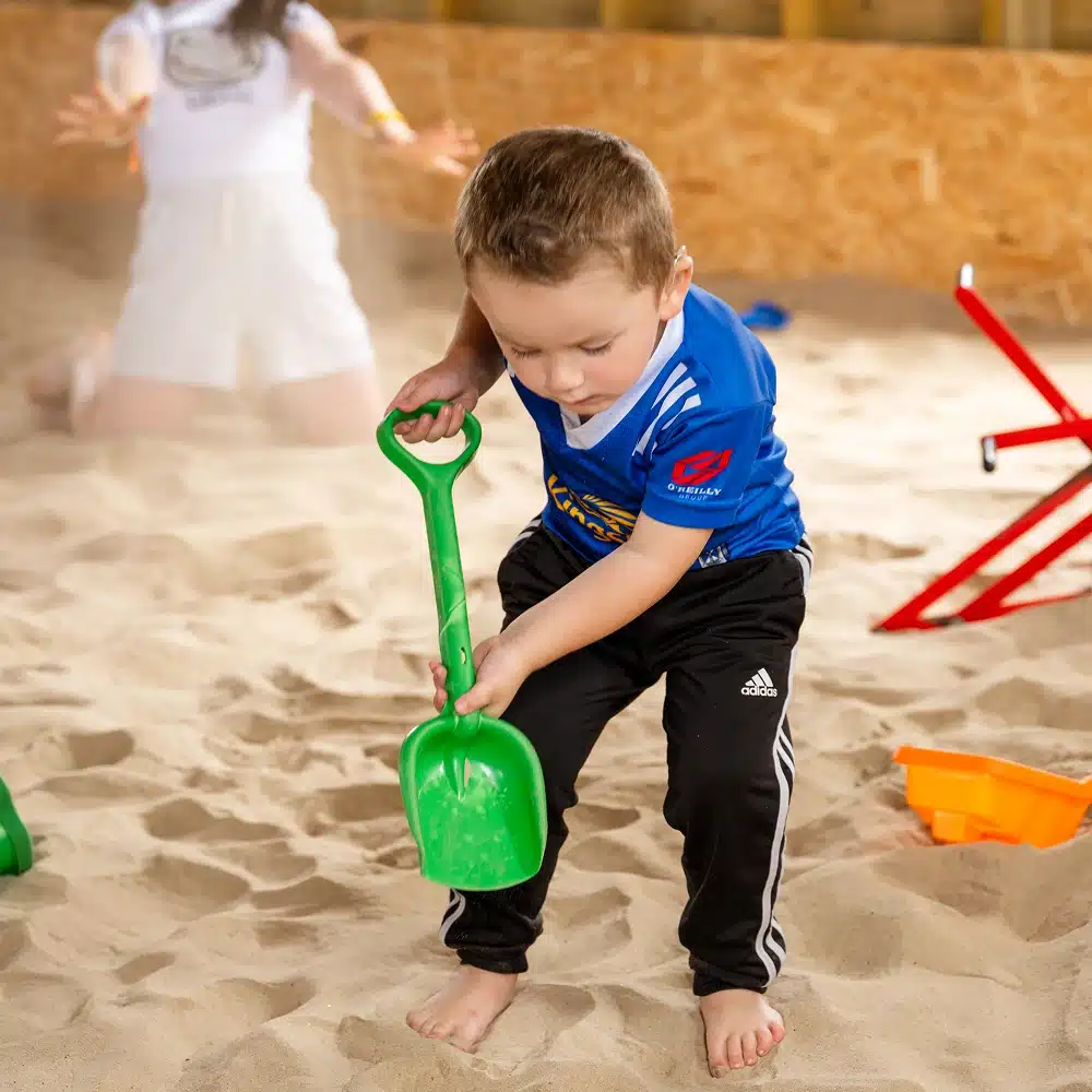 A young boy in a blue shirt and black trousers digs in the sand with a green plastic spade during Navan The Sandbarn activities. In the background, a person in white is blurred, with red and orange toys nearby.