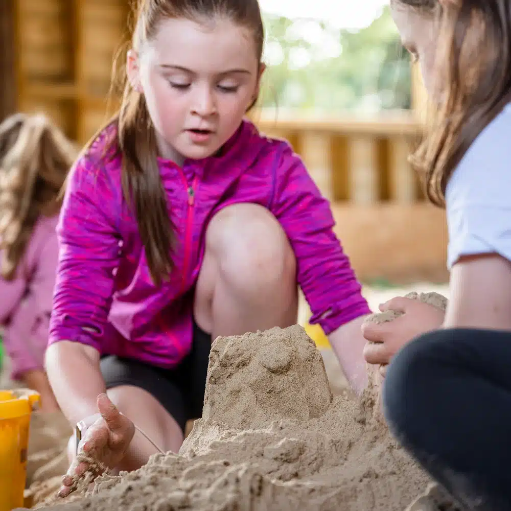 A girl in a pink jacket builds a sandcastle with another child during Navan The Sandbarn activities. They are focused and using their hands to shape the sand, with a yellow bucket nearby. Another child is blurred in the background.