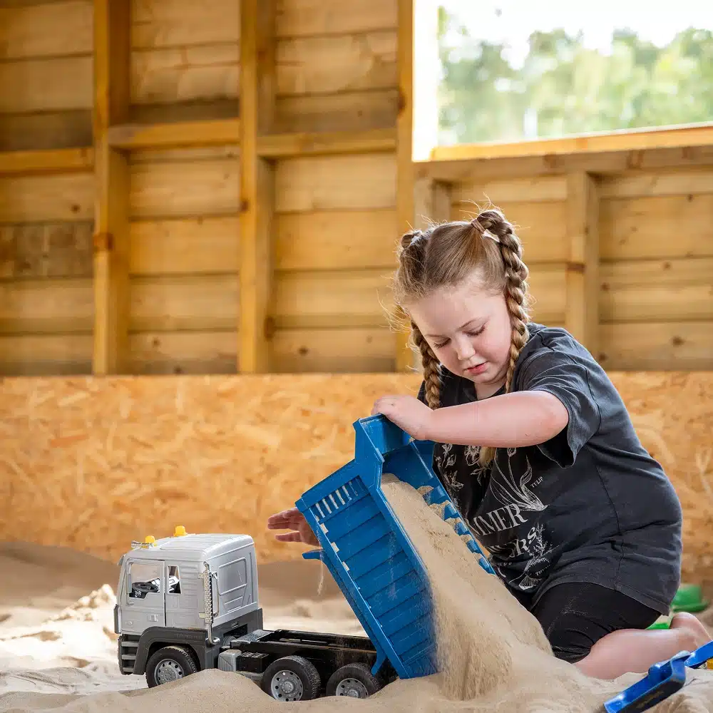 A young girl with plaited hair enjoys Navan The Sandbarn activities indoors, pouring sand from a blue toy dumper truck onto the floor, with wooden walls and an open window in the background.