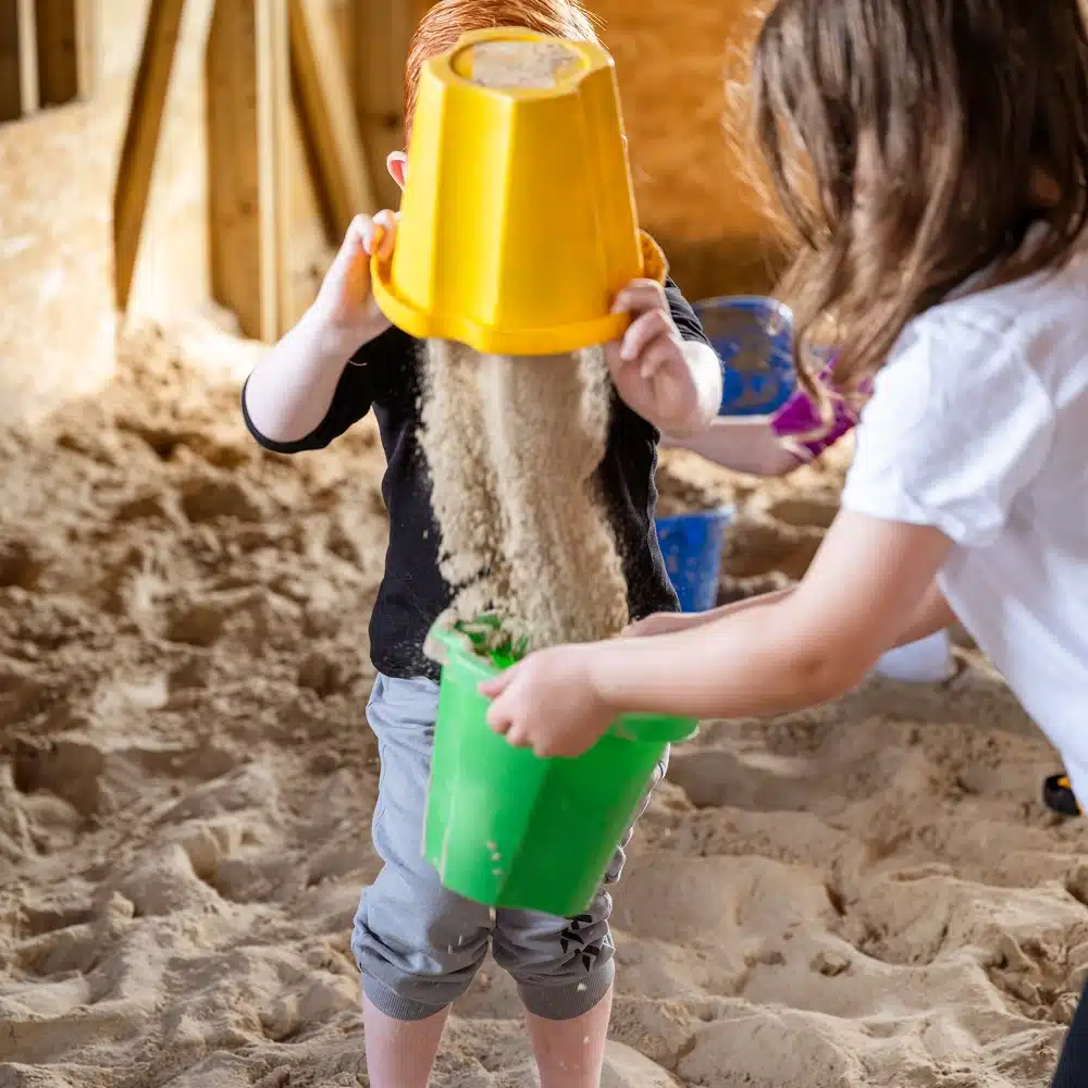 Two children enjoy Navan The Sandbarn activities in a sandy indoor area. One pours sand from a yellow bucket into a green one held by the other. Their faces are mostly hidden, but the scene remains playful and lively in their casual clothes.