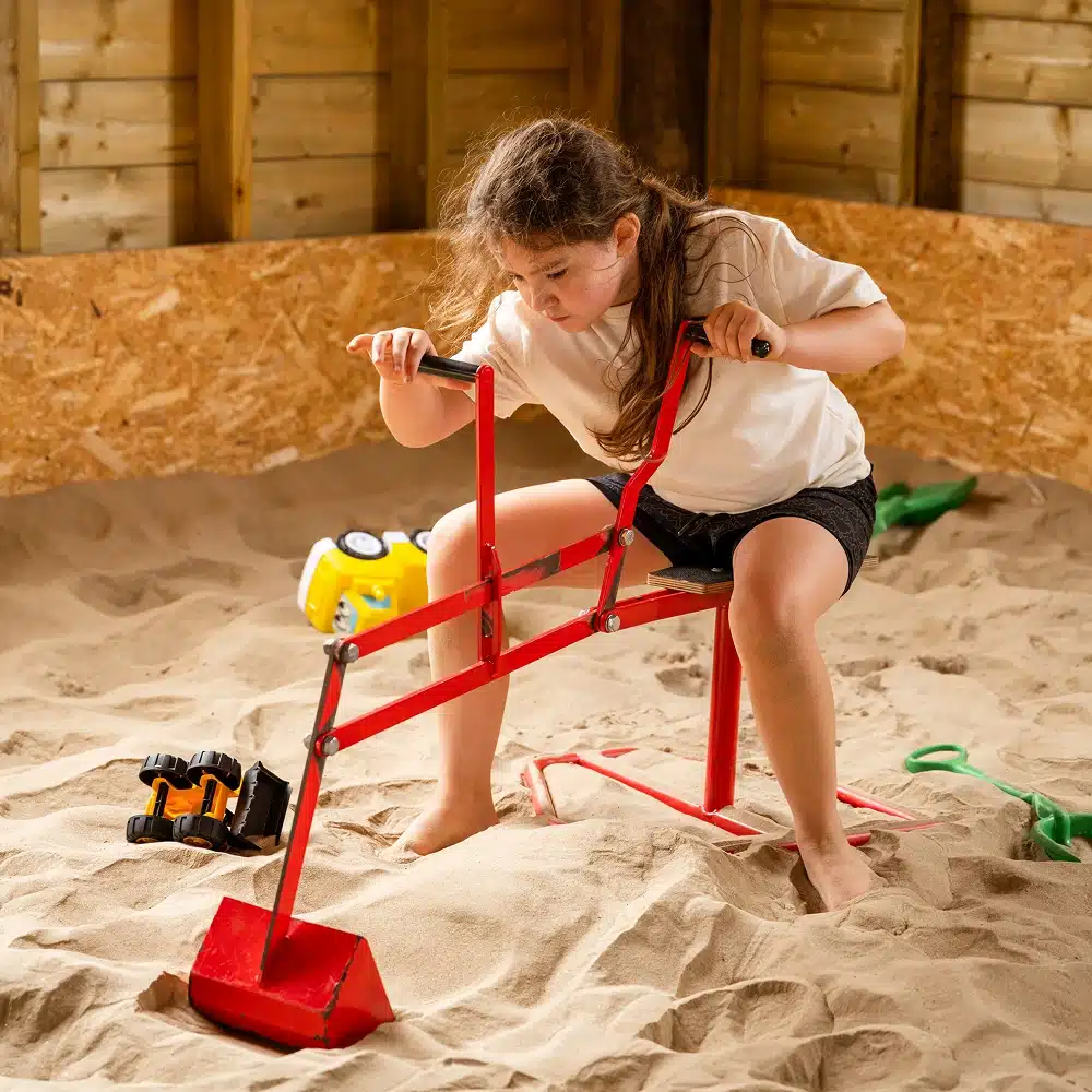 A young girl sits on a red metal digger toy in a sandy indoor play area, using the handles to scoop sand—a perfect example of fun Navan family activities. Toy lorries and a green spade are nearby as she focuses on her task.