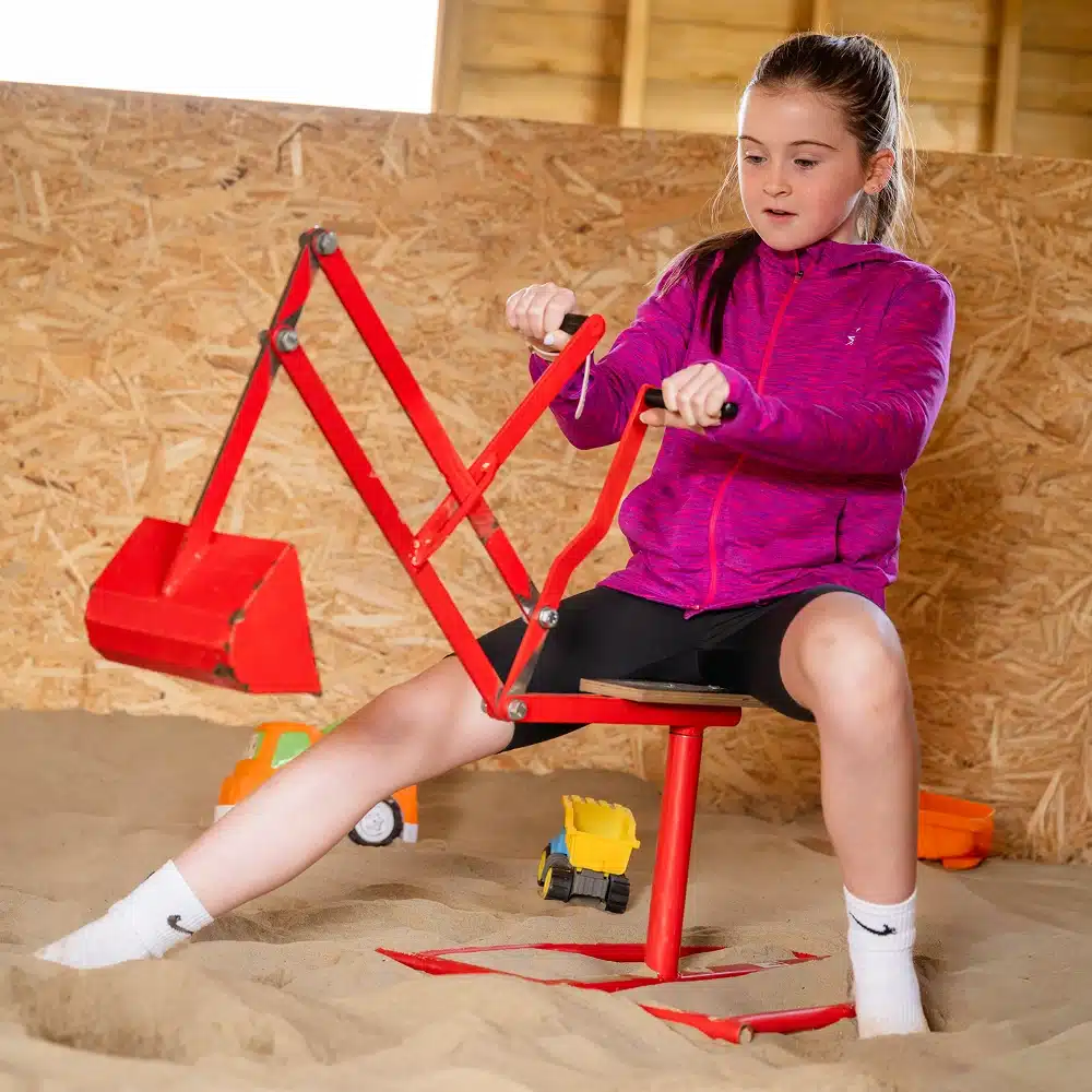 A young girl in a purple jacket operates a red toy digger in the sandy indoor play area at Navan The Sandbarn, enjoying activities surrounded by toy lorries and construction vehicles.