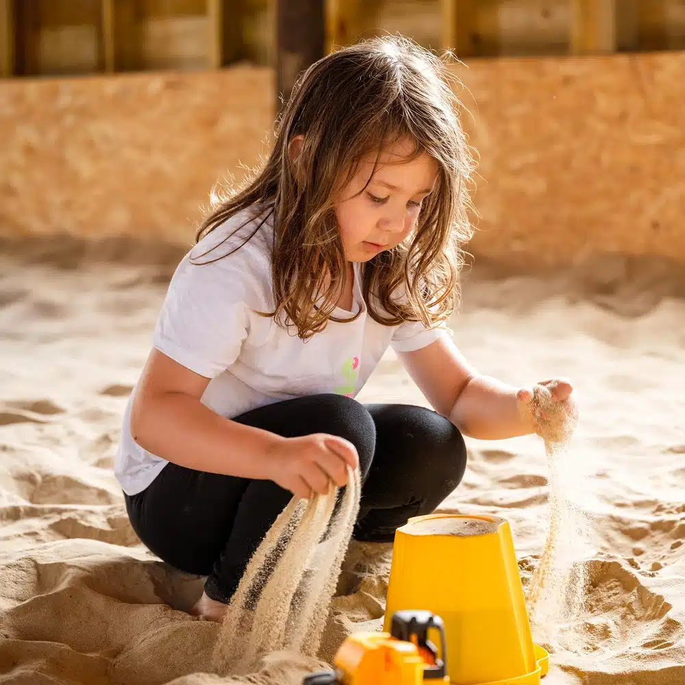 A young girl with long brown hair sits in a sandpit, letting sand pour from her hands into a yellow bucket. She is wearing a white t-shirt and black leggings, enjoying one of the many Navan family activities that spark creativity and focus in children.