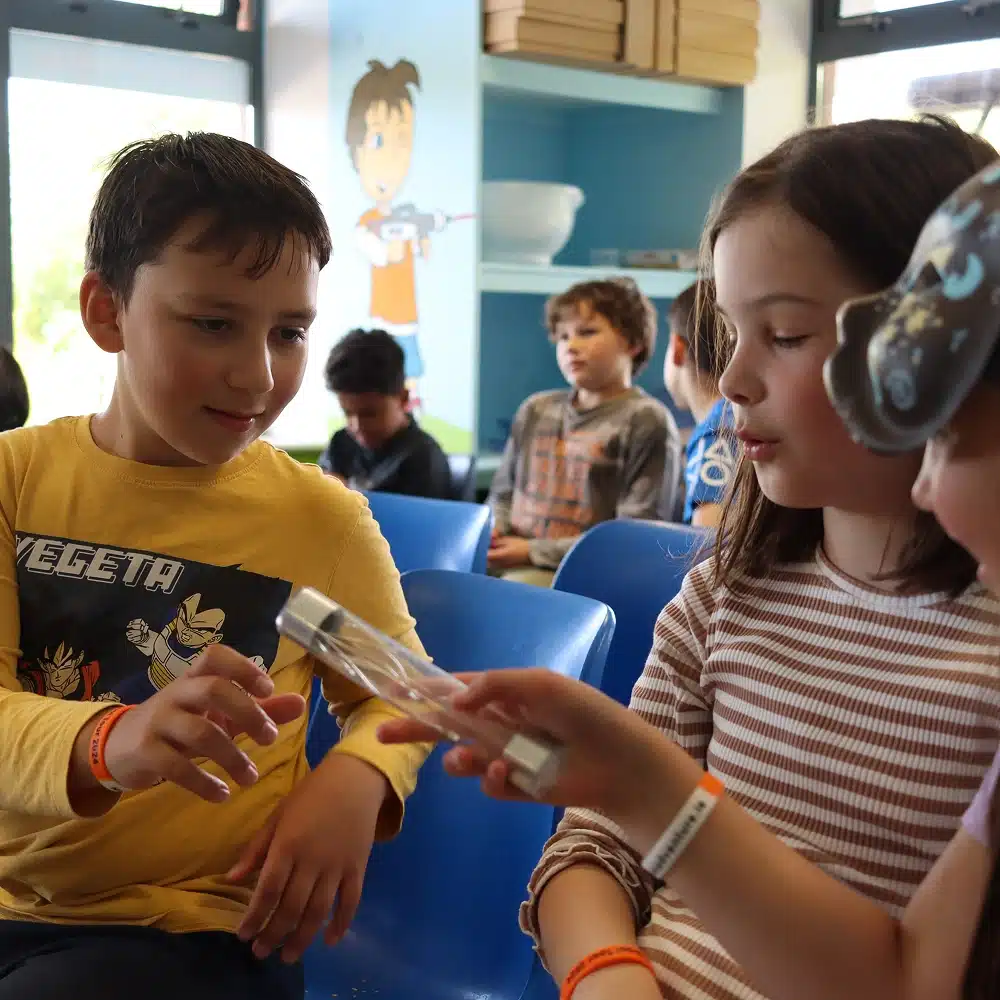 Three children sit on blue chairs, engaging with a science tool—part of a Navan science show for schools; other kids and playful wall art brighten the background of this lively classroom.