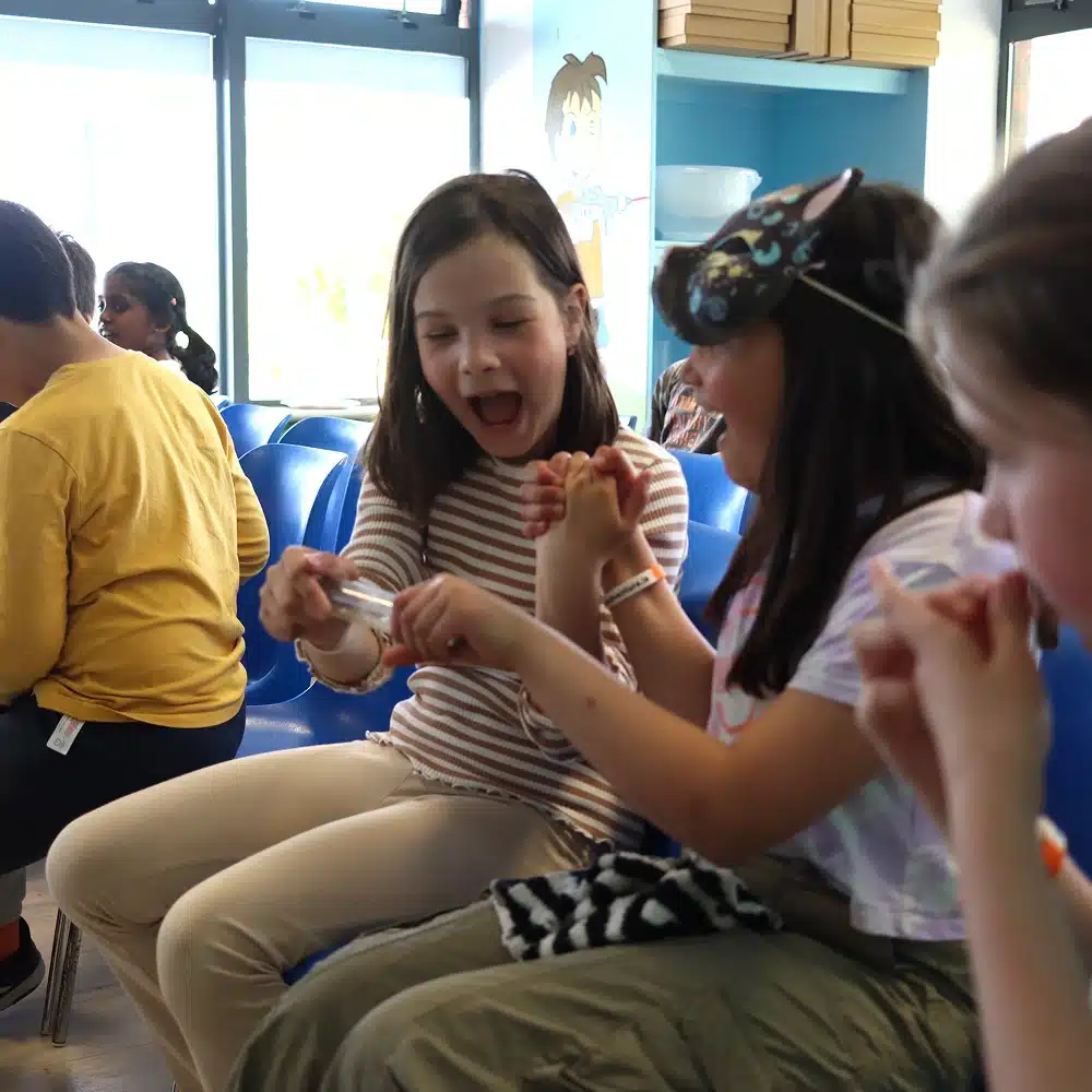 Two young girls sit on blue chairs, smiling and laughing whilst playing with a small object. Another child and an adult are nearby in what appears to be a waiting room or classroom at the Navan science show for schools.