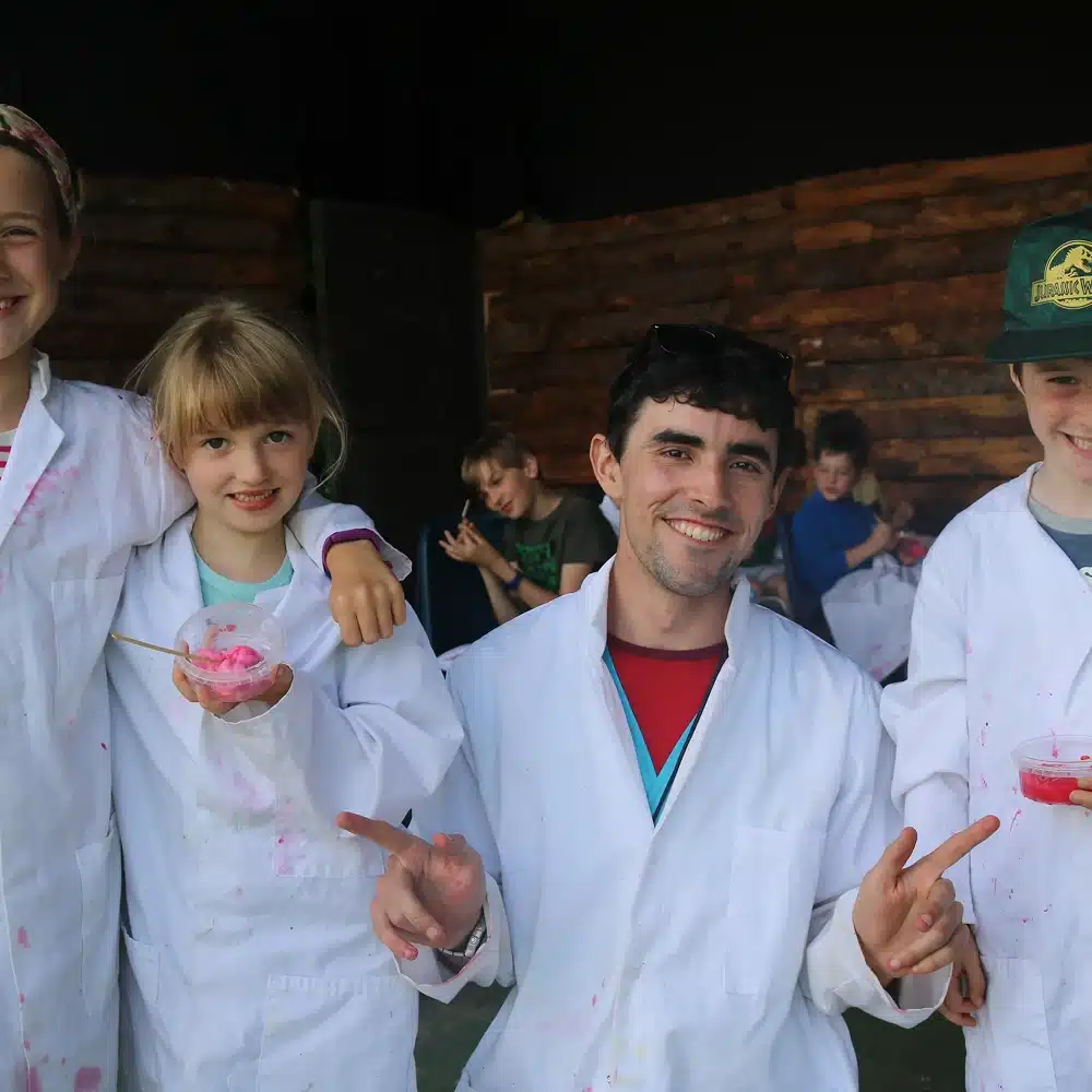 Four children and a young adult in white lab coats smile for the camera, holding bowls with pink substance at a Navan science show for schools, standing in front of a rustic wooden wall. Other children in lab coats are seen in the background.