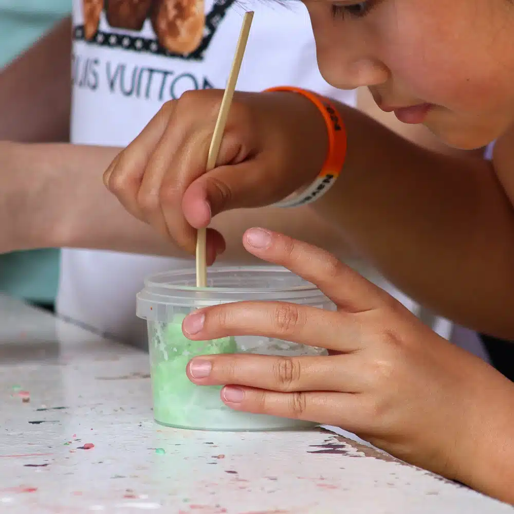 A child stirs a green mixture in a plastic container using a wooden stick, focusing intently on Navan slime making for children. The child wears a bracelet and is seated at a white table with paint splatters.
