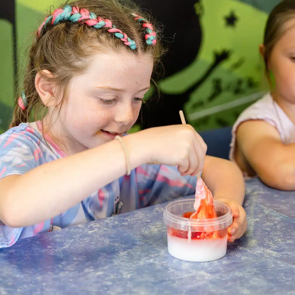 A young girl with plaited hair smiles as she enjoys Navan slime making for children, mixing red slime into a container of white liquid at a table, while another child sits nearby.