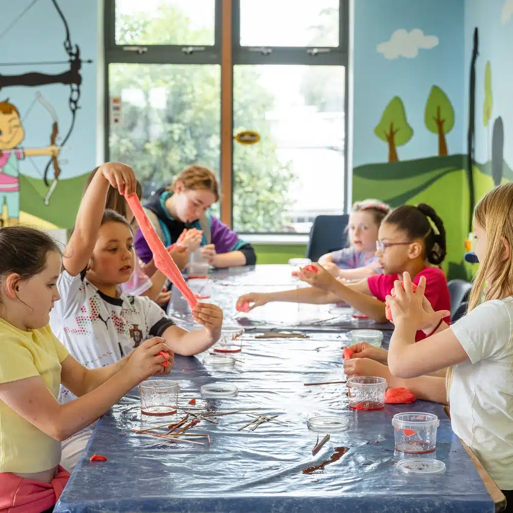A group of young children sit around a table covered with a plastic sheet, enjoying Navan slime making for kids. In the background, a colourful mural shows trees, hills, and a person shooting a bow and arrow.