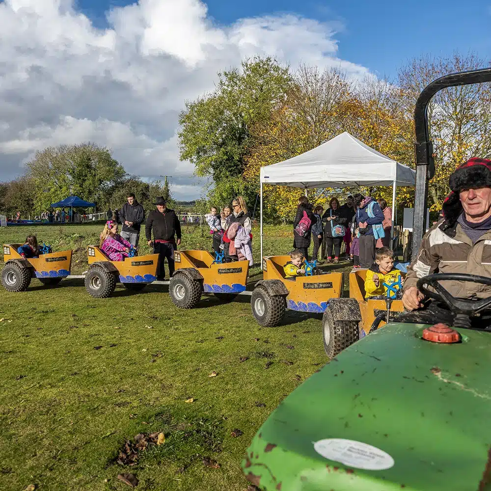Children enjoy a Navan train ride in small yellow carriages pulled by a green tractor at an outdoor event. Adults and children gather under a white marquee, with autumn trees and a partly cloudy sky in the background.