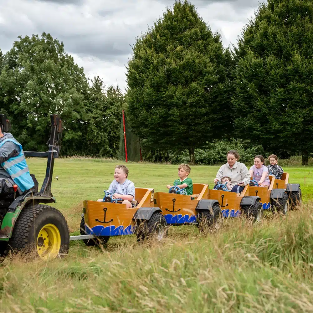 Children and adults enjoy the Navan train ride, travelling in small wooden carriages pulled by a tractor through a grassy field, with trees and cloudy skies in the background.
