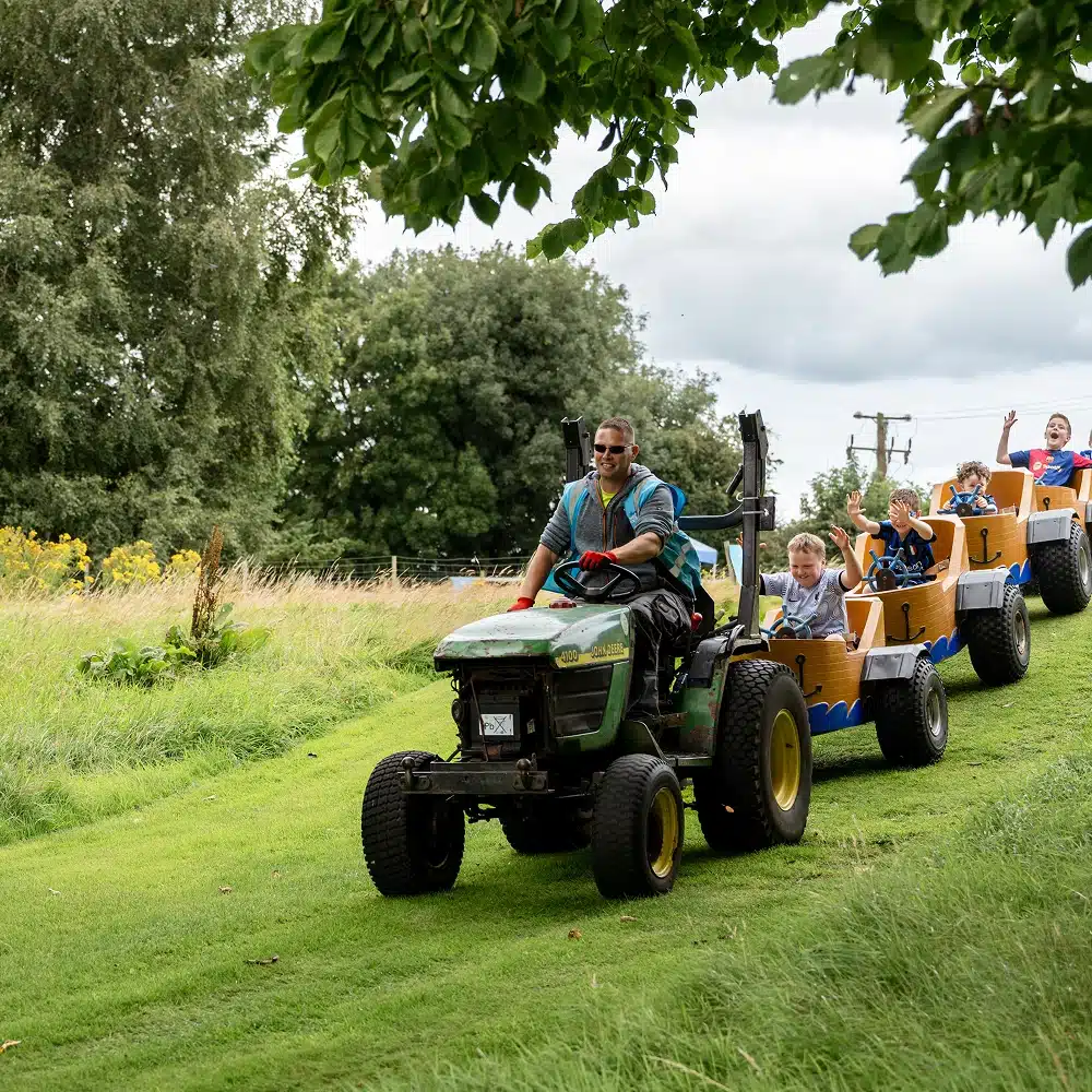 A man drives a small green tractor pulling a train of carts filled with smiling children on the Navan train ride, winding through a grassy, tree-lined outdoor area on a cloudy day.