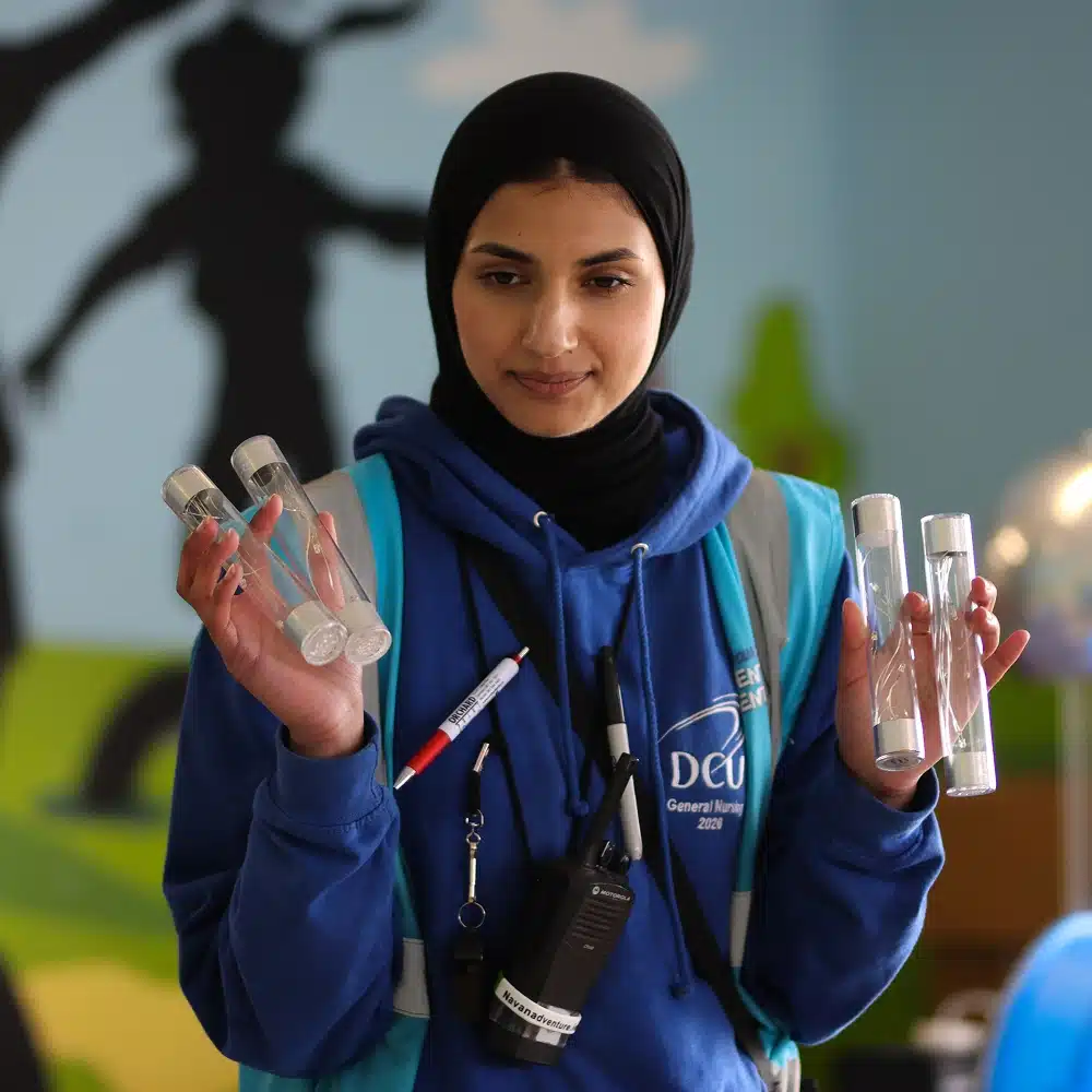 A woman in a blue hoodie and hijab holds four clear tubes, wearing a walkie-talkie and lanyard—perhaps preparing for a Navan bouncy castle party. She stands indoors, with a blurred mural and light blue background behind her.