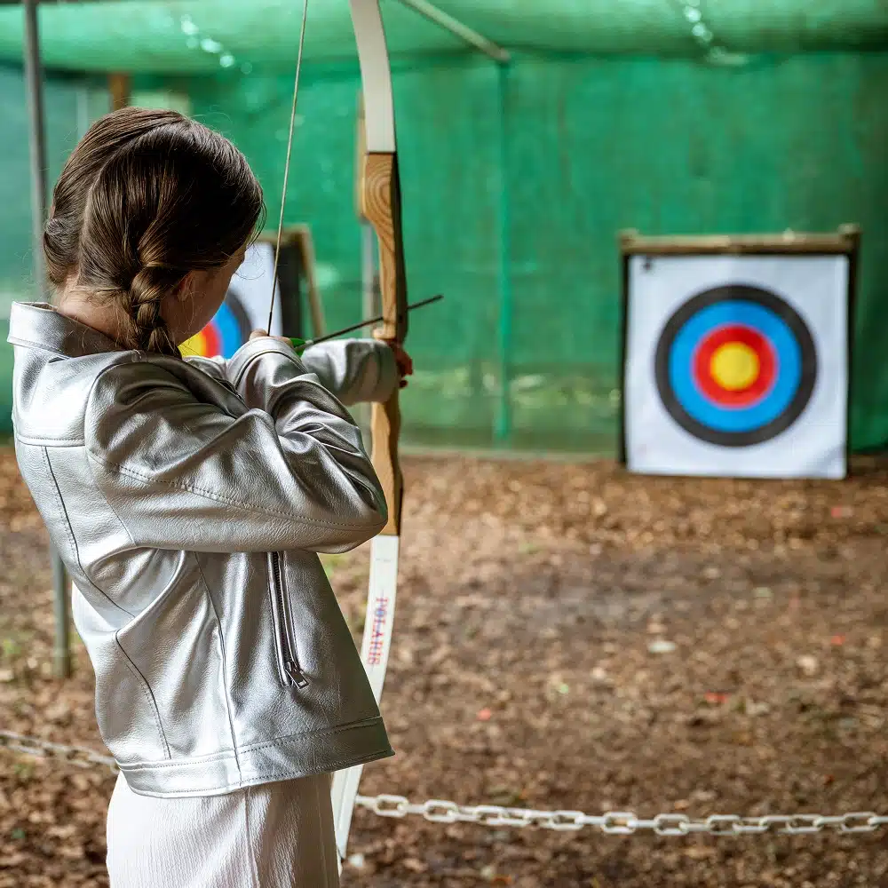 A girl with plaited hair, wearing a shiny silver jacket, aims a bow and arrow at a colourful archery target under a green canopy during a Navan secondary school adventure at the outdoor range.