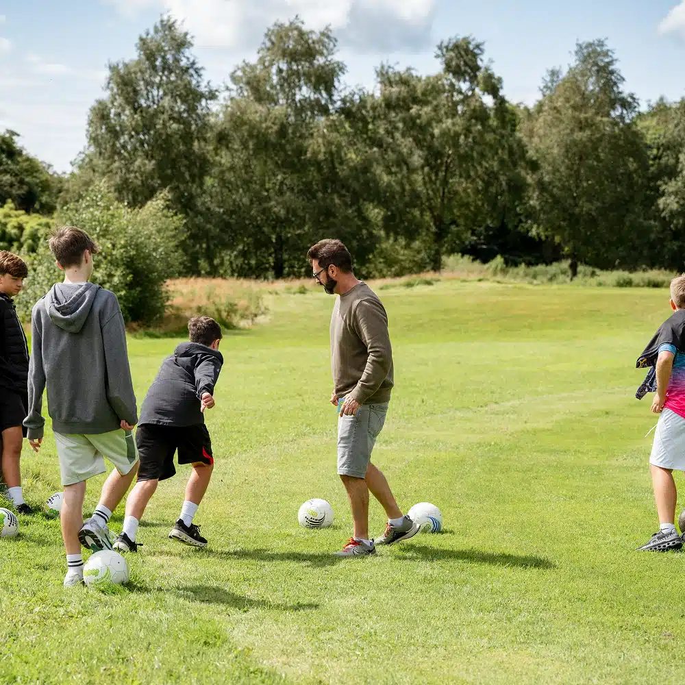 A group of boys and a man play football on a grassy field during a Navan secondary school adventure, with several footballs scattered around and trees in the background under a partly cloudy sky.