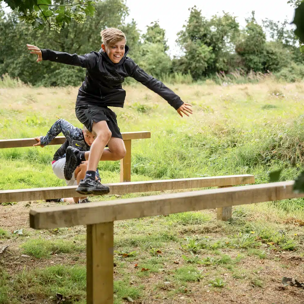 Two children are playing outdoors, jumping over wooden beams in a grassy field—just like a Navan secondary school adventure. The child in front is mid-jump and smiling, while the other lands behind them against a backdrop of lush trees and grass.
