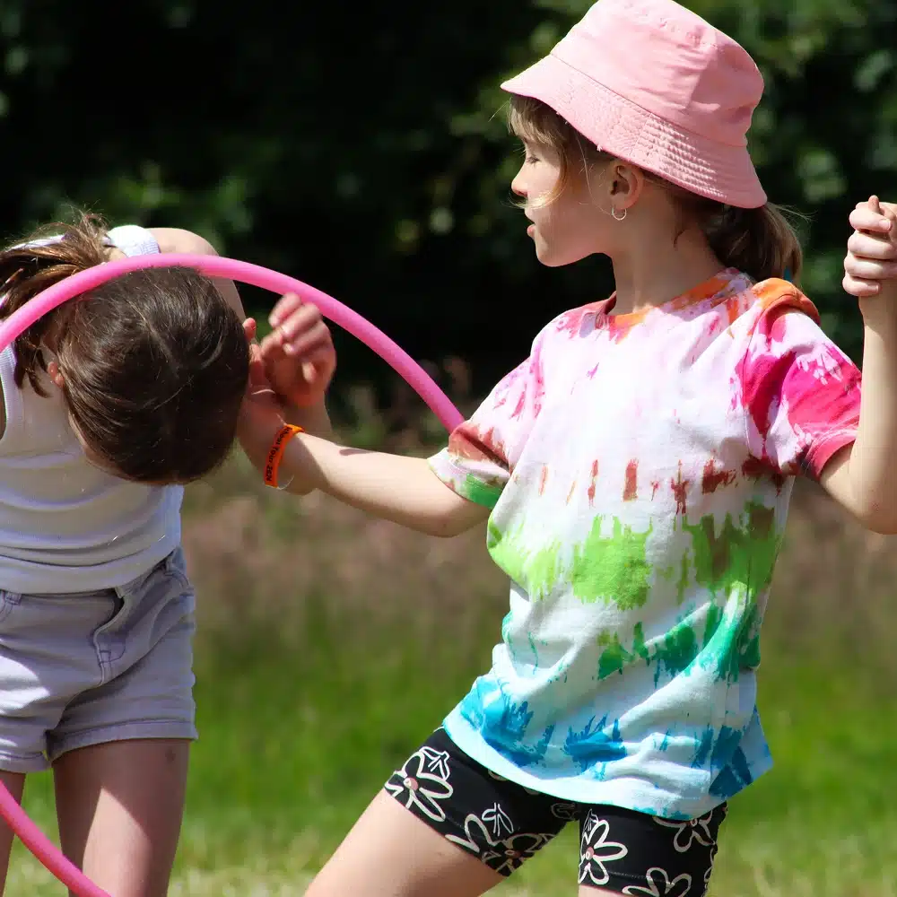 Two children play outside on a sunny day, surrounded by green grass and trees. Like a Navan secondary school adventure, one in a pink hat and tie-dye shirt helps the other as they bend forward through a bright pink hula hoop.