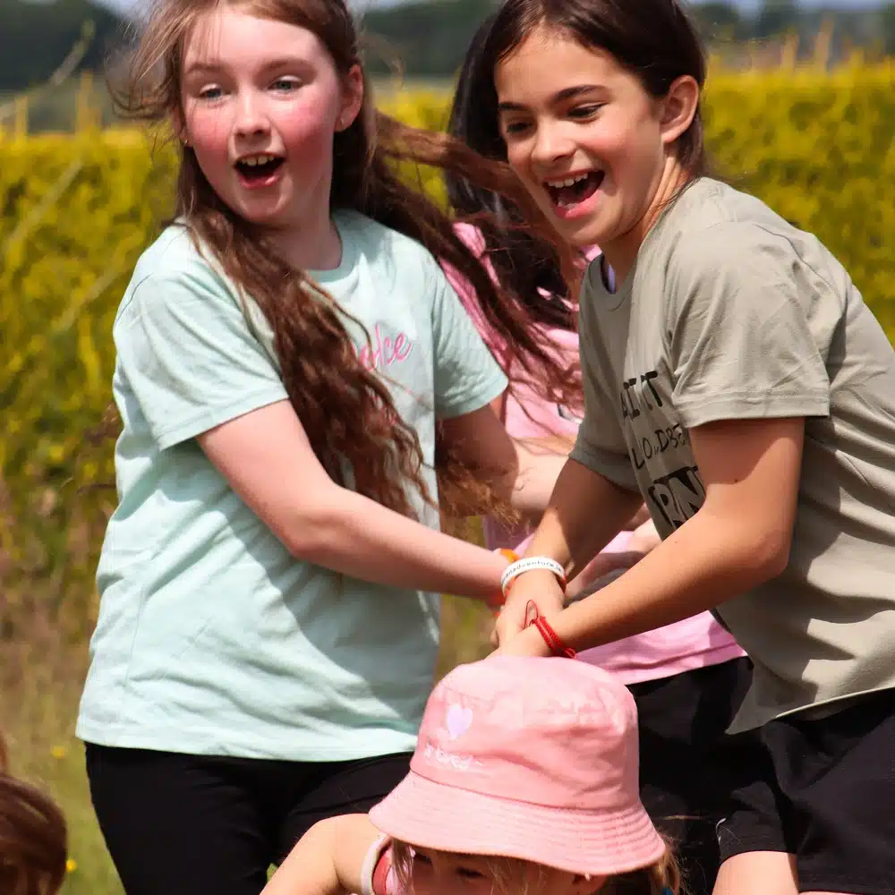Two young girls, smiling and holding hands, enjoy a playful Navan secondary school adventure outdoors on a sunny day. One wears a light blue shirt, the other beige. A child in a pink hat is partially visible in the foreground against green scenery.