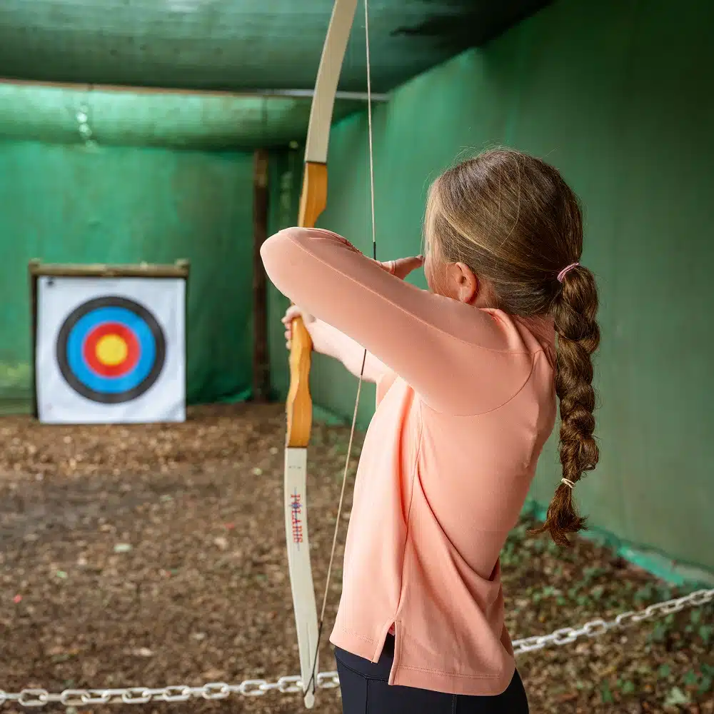 A girl with a long plait, wearing a peach long-sleeved shirt, aims a bow and arrow at a colourful target indoors during a Navan primary school adventure, standing confidently behind a chain barrier.