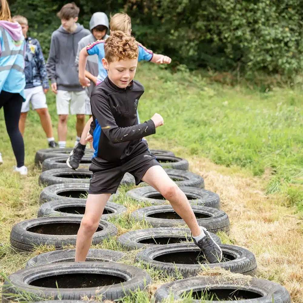 A young boy in sports clothes steps through a tyre obstacle course outdoors at a Navan primary school adventure, with other children and an adult watching in the background on a grassy field.