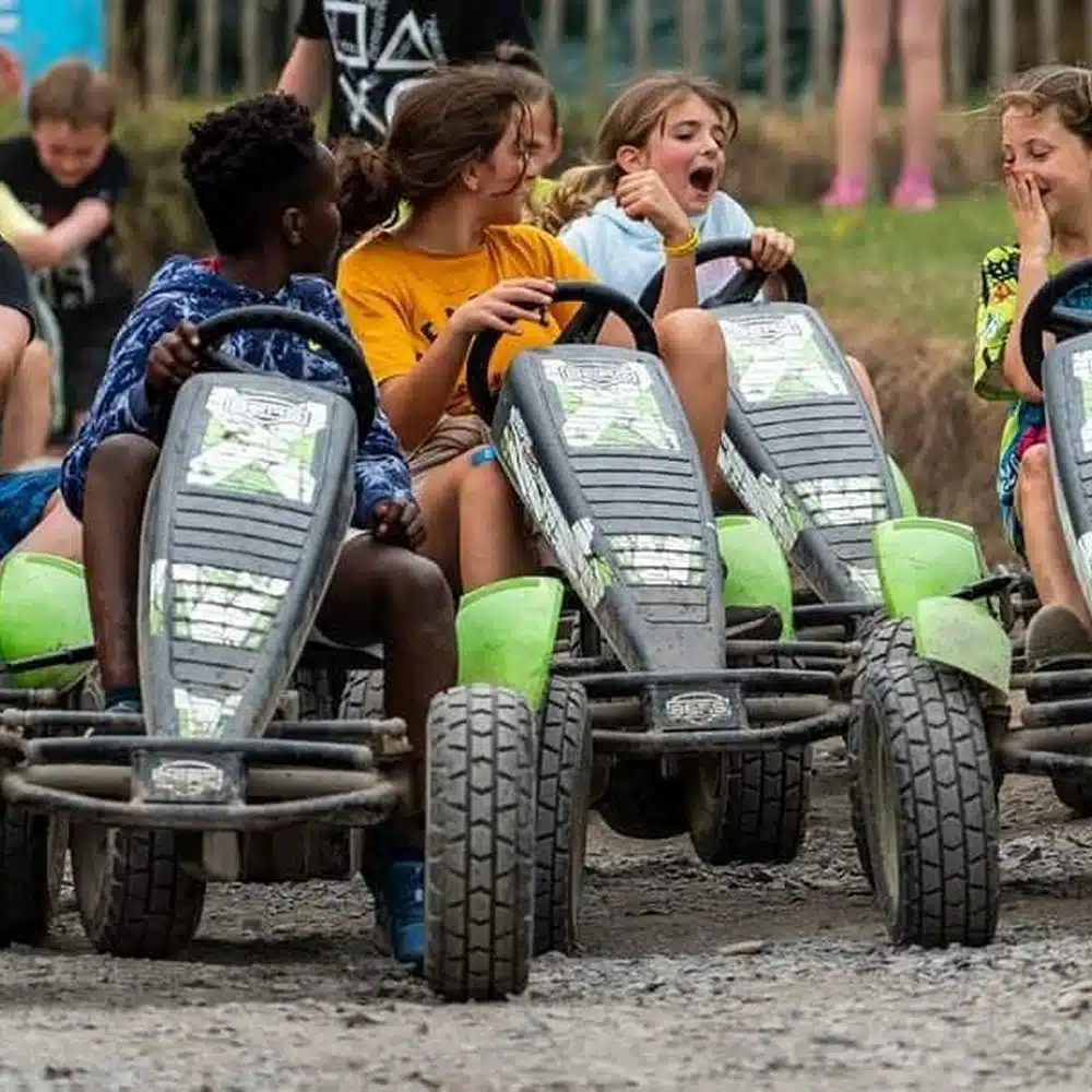 Four children sit in green and black pedal go-karts on a dirt track, smiling and laughing together during a Navan primary school adventure. Other kids are visible in the background, watching and enjoying the activity.