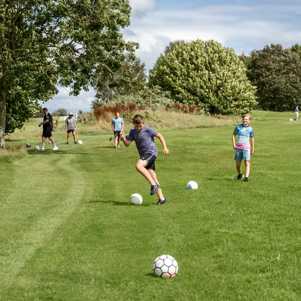 Five children from Navan primary school are playing football on a grassy field surrounded by trees. One child is about to kick the ball, while others watch or walk in the background, enjoying their adventure under a partly cloudy sky.