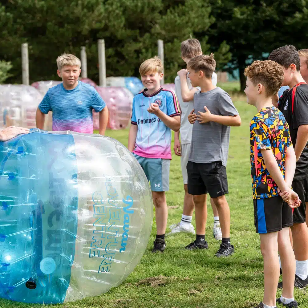 A group of boys stand on grass, smiling and laughing around a person inside a large inflatable zorb at a Navan bubble football party. More zorbs are visible in the background, with trees and wooden posts nearby.