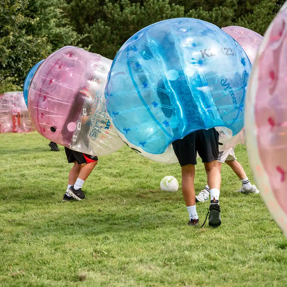 People enjoying a Navan bubble football party outdoors on grass, each wearing large inflatable bubbles over their upper bodies. Only their legs are visible as they chase a football, with trees in the background.