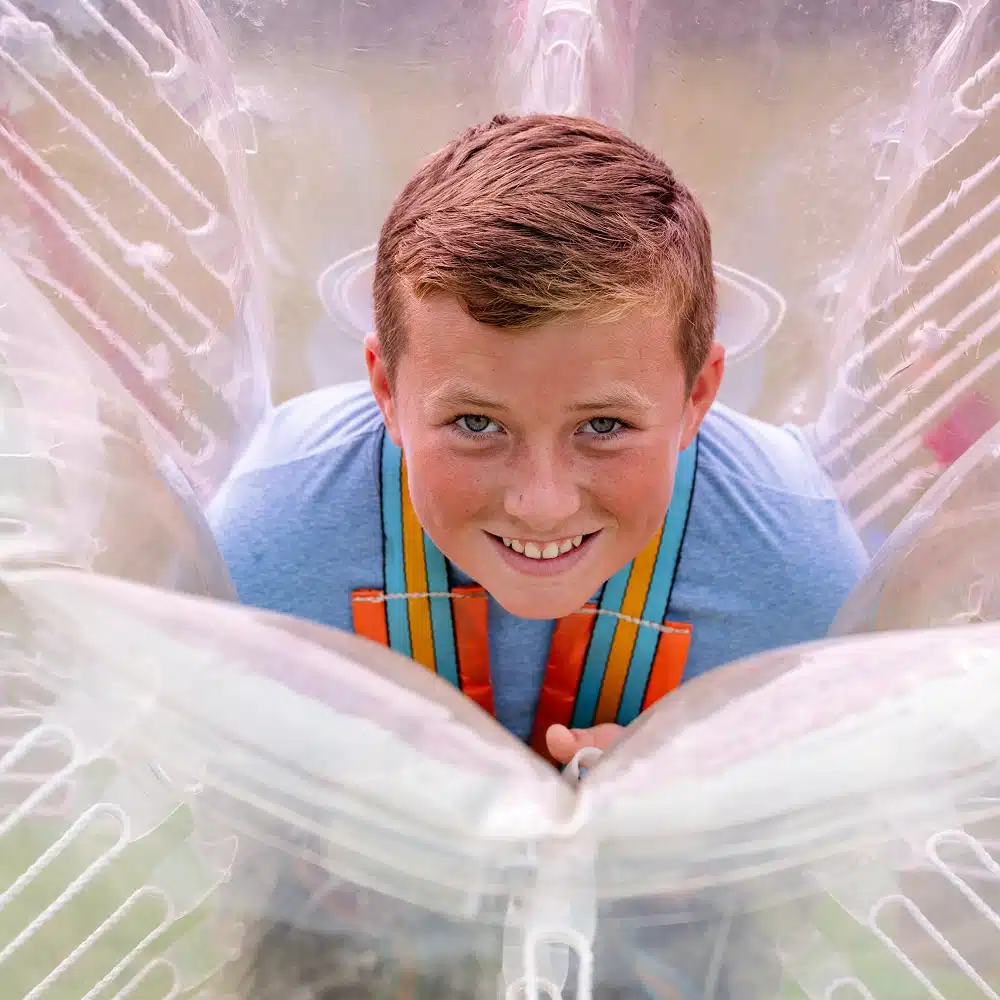 A smiling boy with short brown hair looks up whilst inside a large, clear inflatable bubble suit at a Navan bubble football party, wearing a blue shirt with multicoloured straps over his shoulders.