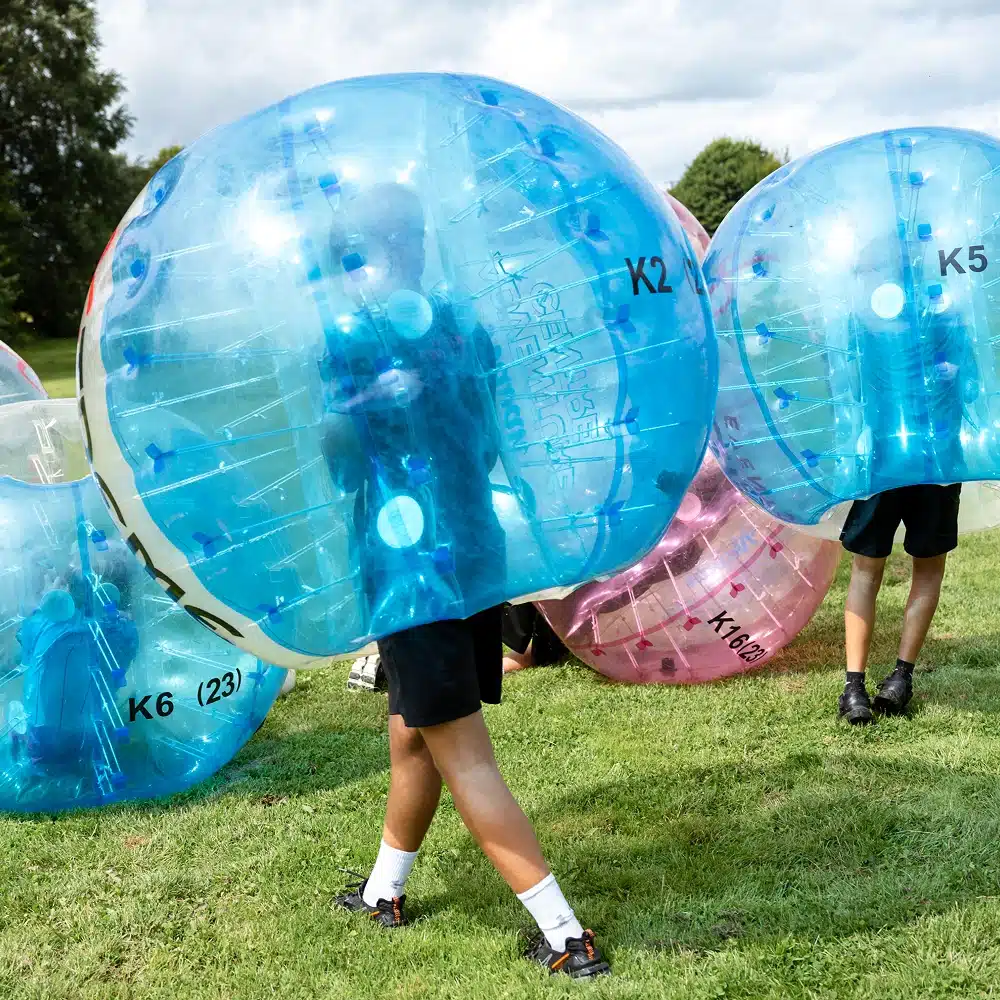 People enjoying a Navan bubble football party on a grassy field, each wearing large inflatable bubbles over their upper bodies and heads. Only their legs are visible as they walk or run inside the bubbles.
