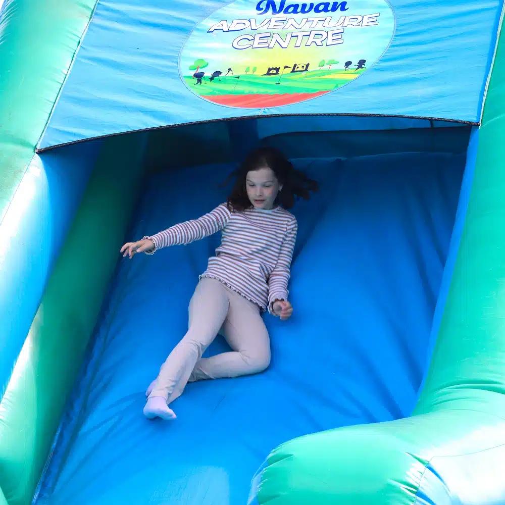 A young girl in light-coloured trousers and a striped top slides down a blue inflatable slide at Navan Adventure Centre, where Navan bubble football party activities bring extra excitement. She looks focused as she descends, hands raised for balance.