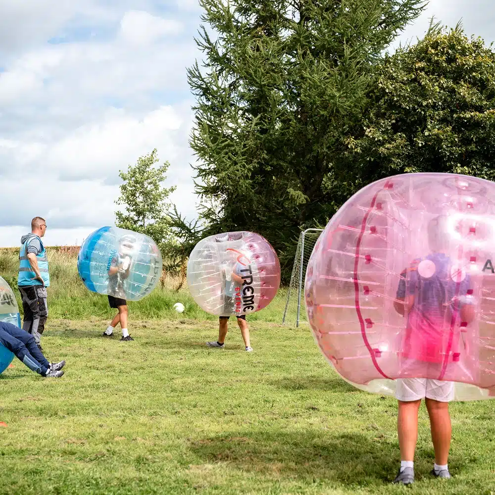 People enjoying a Navan bubble football party outdoors on grass, each wearing large inflatable bubbles—some clear, some pink. Trees and a small football goal are set against a partly cloudy sky in the background.