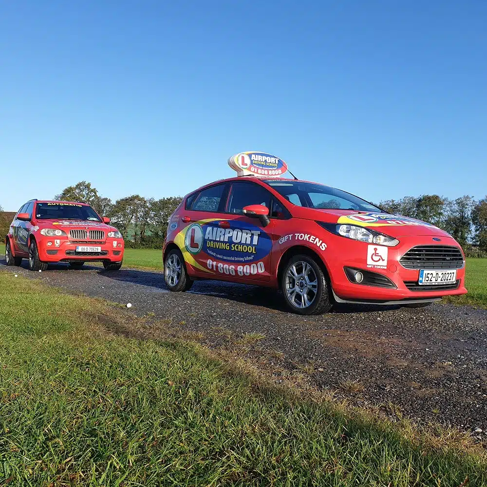 Two red cars with "Airport Driving School" logos are parked on a rural road. Displaying L-plates, wheelchair accessibility symbols, and Navan junior driving programme details, they sit amid green grass and trees beneath a clear blue sky.