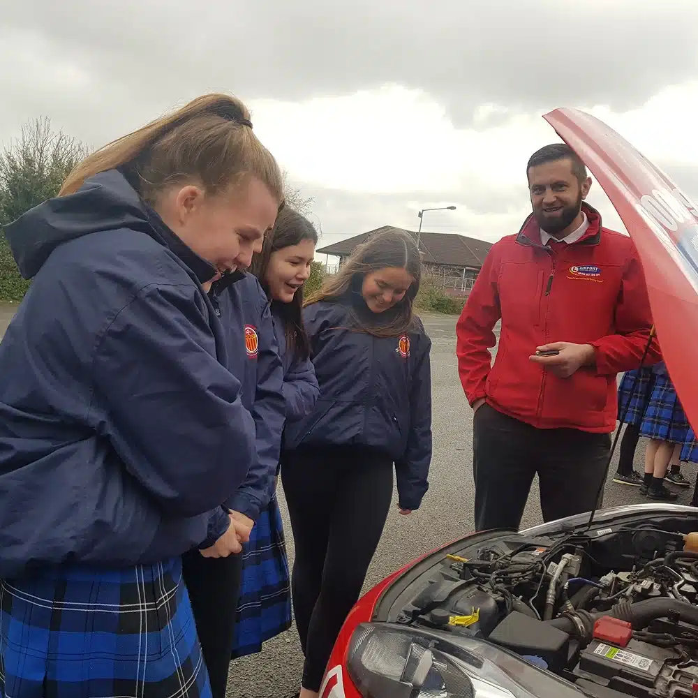 Three girls in blue uniforms and a man in a red jacket stand outdoors, smiling at the open bonnet of a red car as part of the Navan junior driving programme, observing the engine together on a cloudy day.