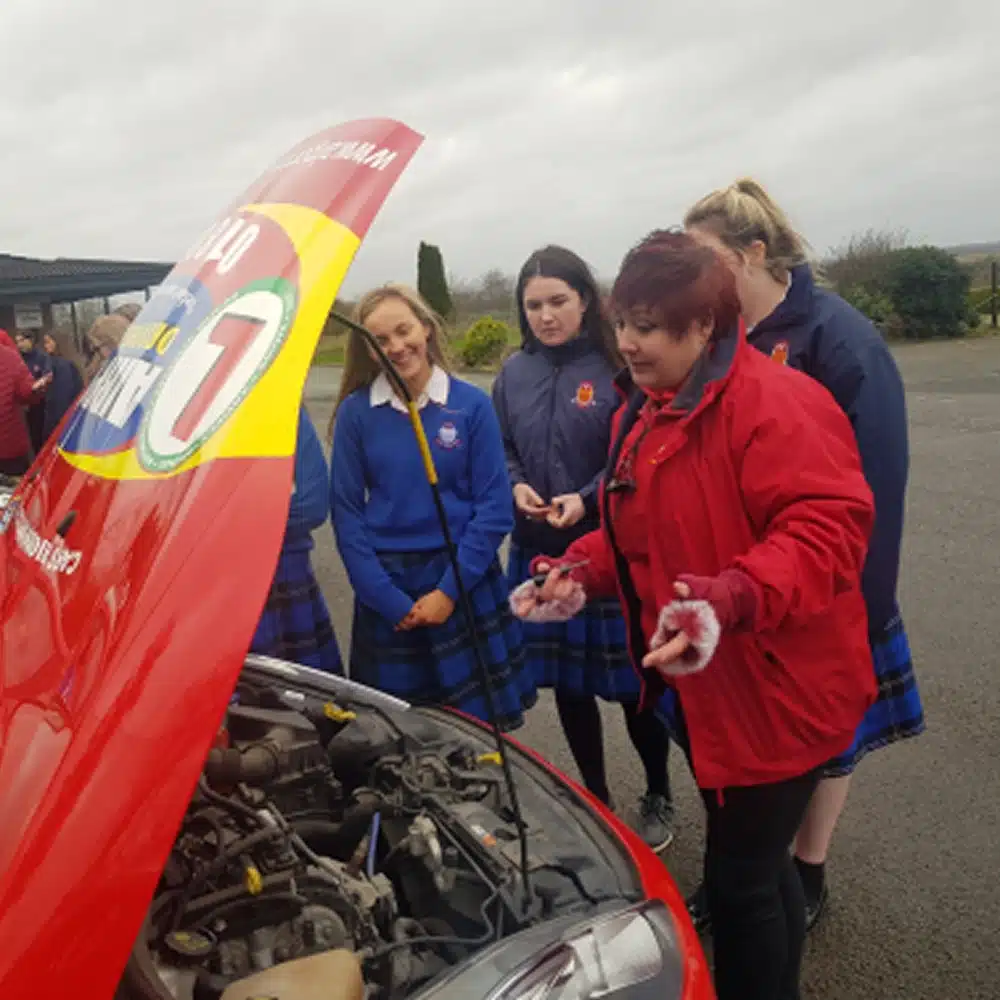 A group of schoolgirls in uniforms observe as a woman in a red jacket explains something under the open bonnet of a red car, part of the Navan junior driving programme, on a cloudy day.