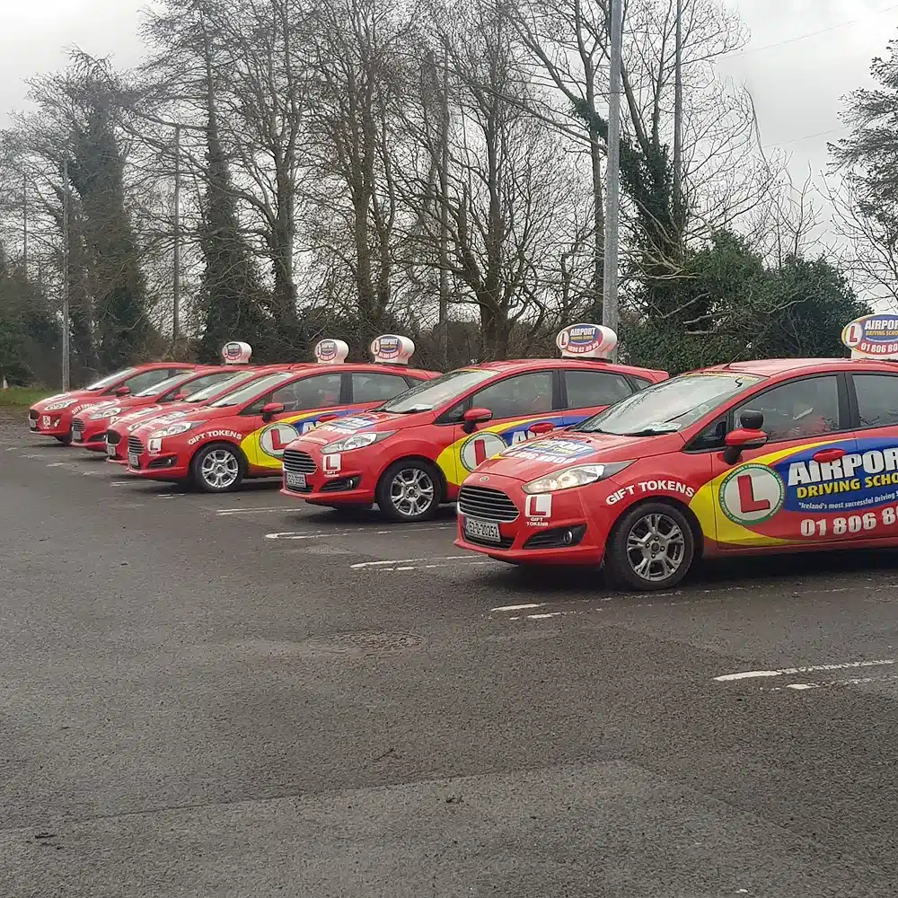 A row of red cars with "Airport Driving School" signs and L-plates, part of the Navan junior driving programme, are parked in a car park on a cloudy day, with bare trees and greenery in the background.