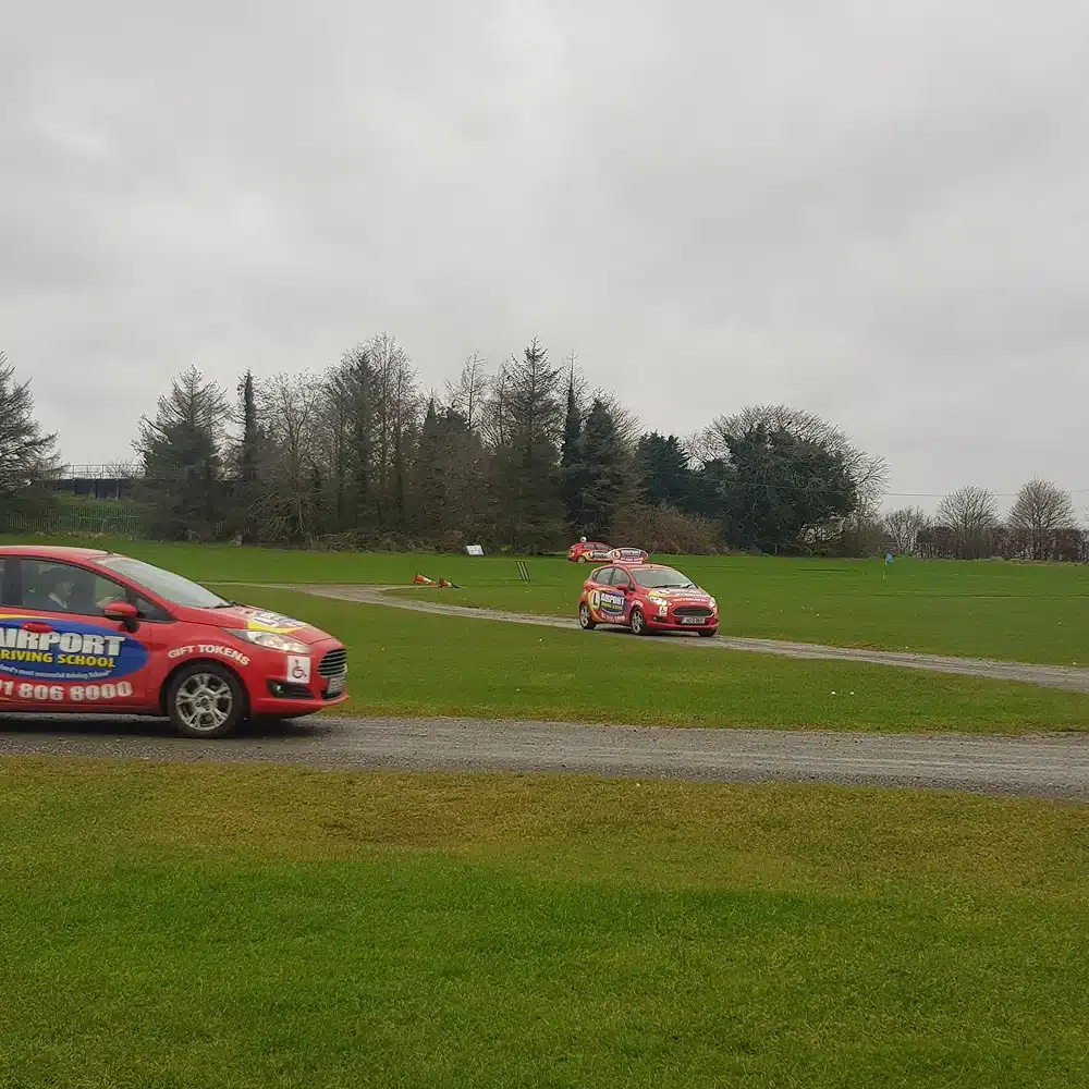 Two red cars with colourful Navan junior driving programme advertisements are parked on a grassy field near a narrow paved path, with trees and an overcast sky in the background.