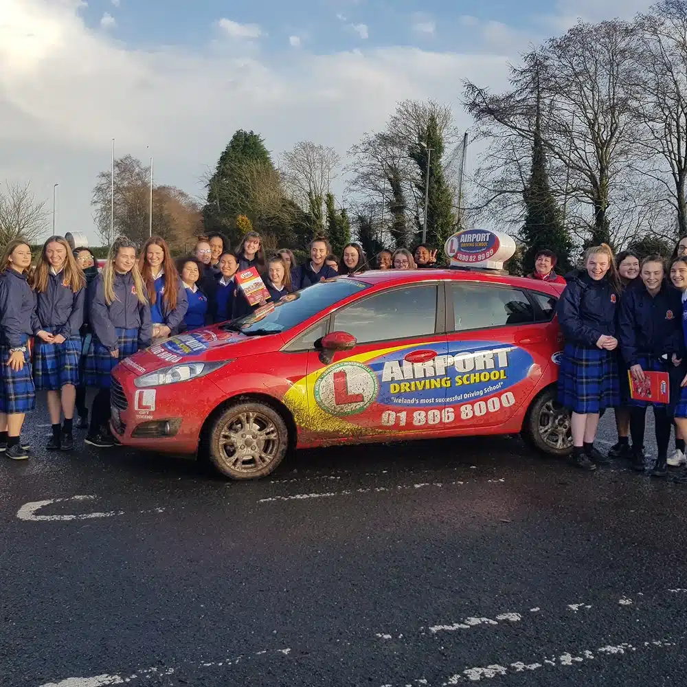 A group of students in blue tops and plaid skirts stand around a red Airport Driving School car on a wet road, smiling for the camera, as part of the Navan junior driving programme, with trees and a cloudy sky in the background.